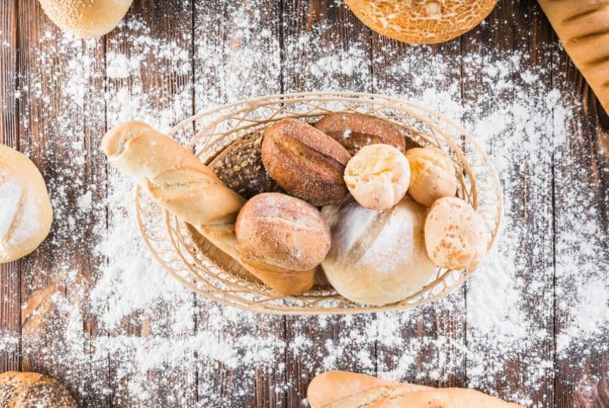 На столі з розсипаним борошном стоїть кошик з хлібом та пампушками На столі з розсипаним борошном стоїть кошик з хлібом та пампушками